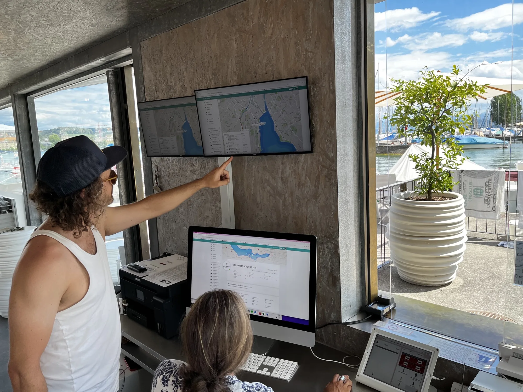 Two staff at a boat rental counter on Lake Zurich: the coride fleet map runs on two wall-mounted screens while a hull's detail view is open on the desktop, with the marina visible through the window